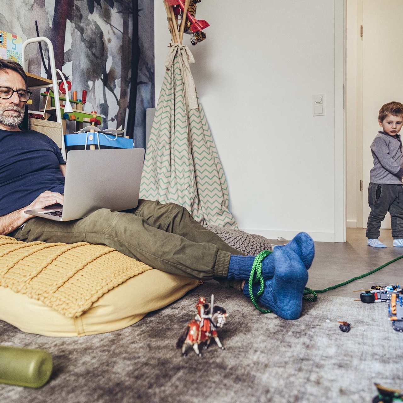 Der Vater macht gerade Home Office und der kleine Junge hat ihn mit einem Seil die Beine gebunden